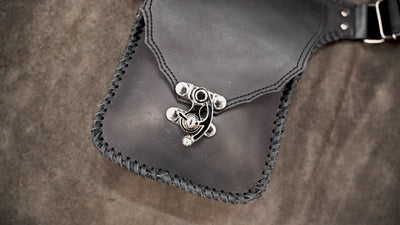 Handmade black leather thigh bag with adjustable waist strap on a textured background.