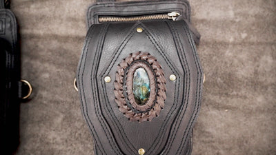 Artisan-crafted black leather shoulder holster featuring dual zip pockets, brass rings, and stone accents.